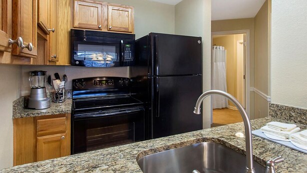 Una cocina con armarios de madera, un microondas negro, un refrigerador negro, un lavamanos de acero inoxidable y una encimera de granito.
