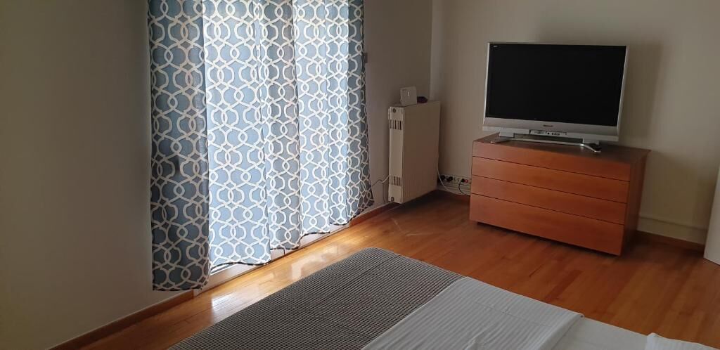 Un dormitorio con cama, televisor sobre una cómoda, radiador y cortinas estampadas.