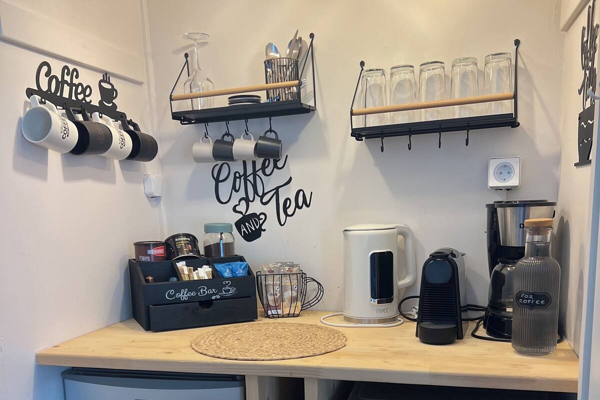 A compact kitchen with a coffee station, a refrigerator, a dishwasher, and a small trash bin.