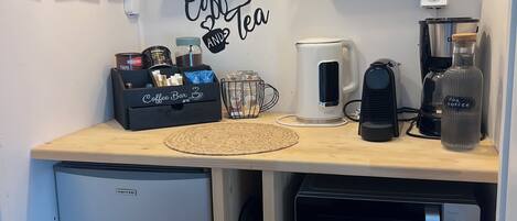 A compact kitchen with a coffee station, a refrigerator, a dishwasher, and a small trash bin.
