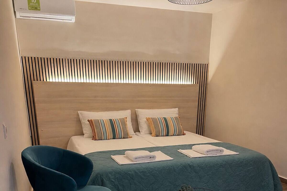 A modern hotel room with a bed, two blue chairs, a wooden coffee table, and a ceiling light fixture.