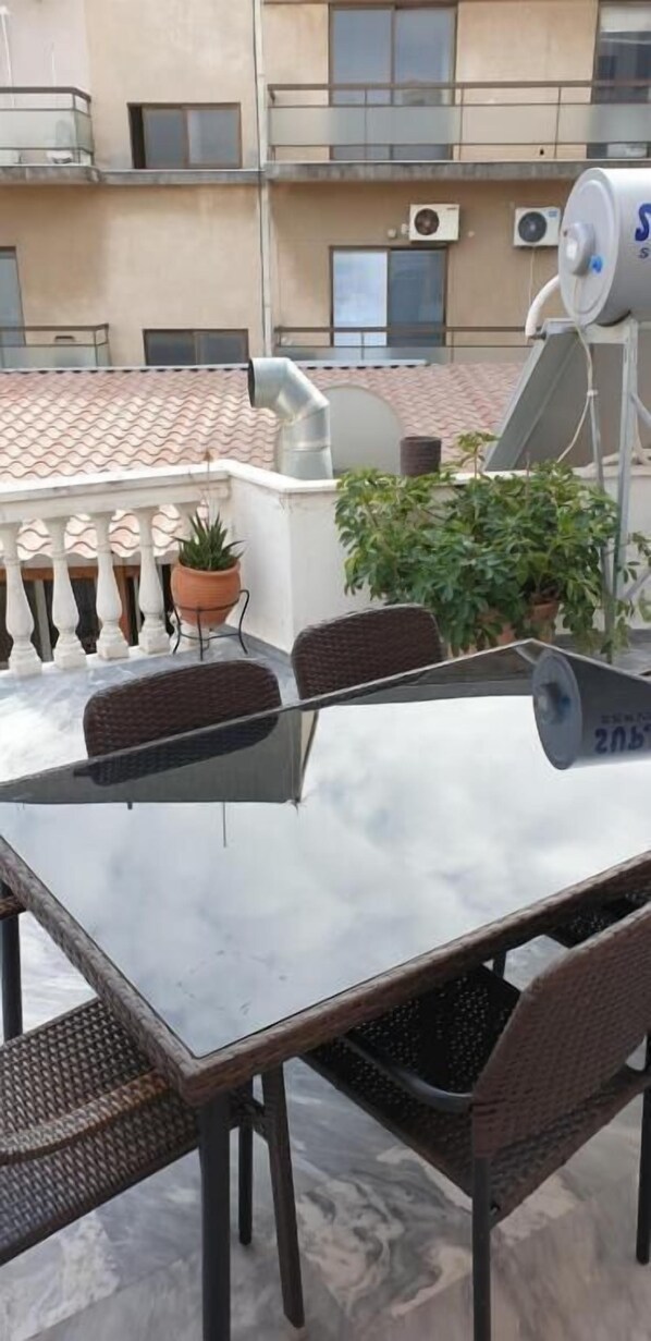 Una terraza en la azotea con una mesa de vidrio y sillas de mimbre, rodeada de plantas en macetas.