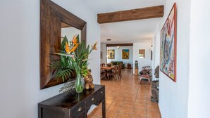 Un couloir avec une console en bois, un miroir et un vase avec des plantes. Au fond, on aperçoit un coin repas avec une table et des chaises.