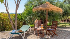 Un espace extérieur ensoleillé, aménagé avec des sièges, comprenant un parasol en chaume, deux chaises en bois, une table avec une boisson, et une chaise longue avec une serviette et un chapeau.