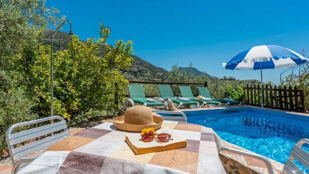 A poolside area with a checkered table, a straw hat, sunglasses, and a book. There are lounge chairs and a blue and white umbrella.
