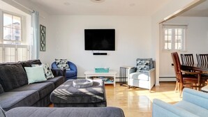 Una sala moderna con un televisor de pantalla plana montado en la pared, un sofá gris, un sillón azul y una zona de comedor con mesa y sillas de madera.