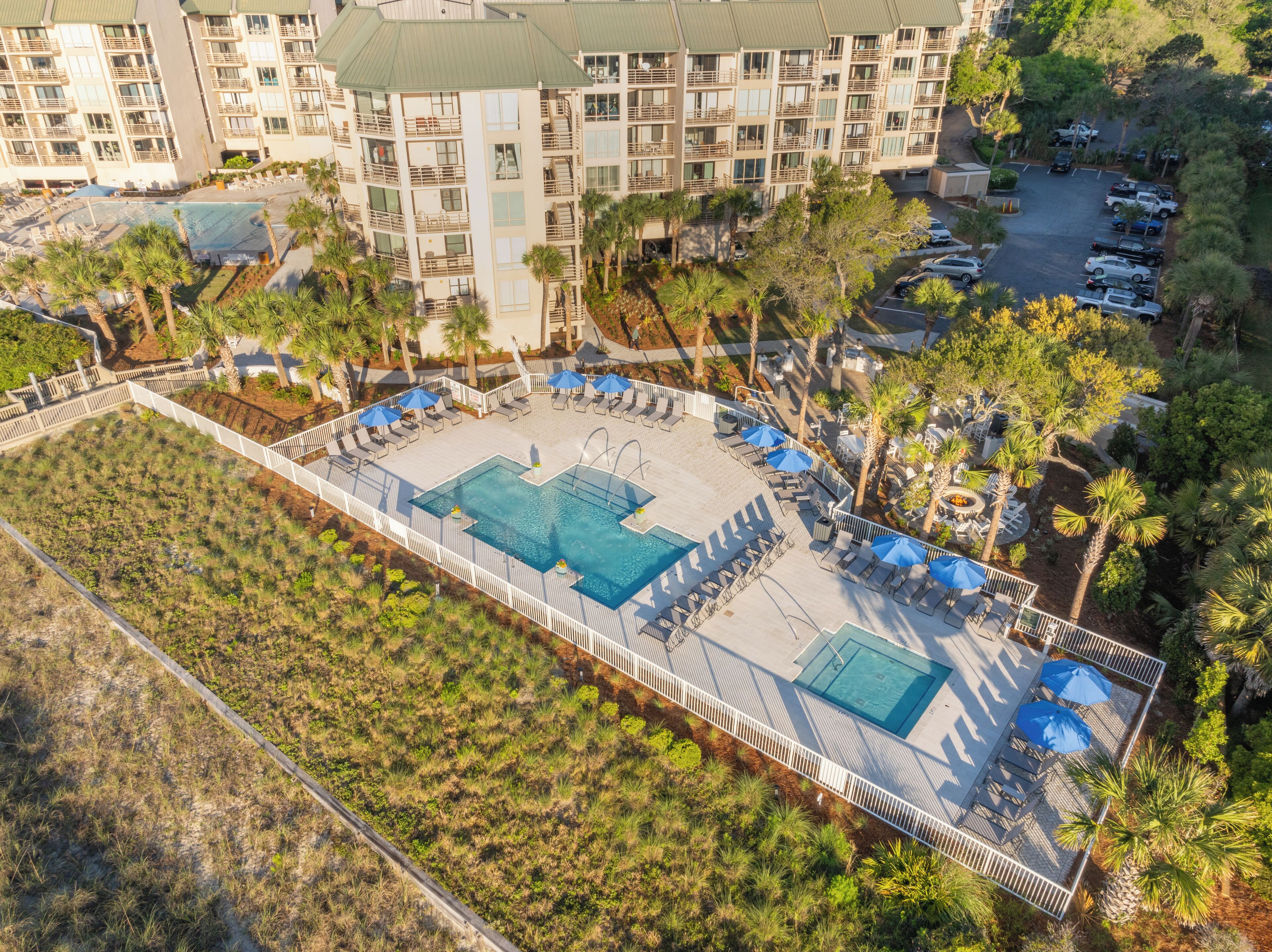 An aerial view of a hotel with a central swimming pool surrounded by lounge chairs and umbrellas, with a parking area and multi-story buildings in the background.