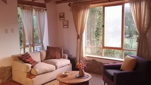 Ein gemütliches Wohnzimmer mit einem beigen Sofa, einem violetten Sessel und einem Holztisch. Zwei Fenster mit Vorhängen, eines davon bietet Blick ins Grüne.