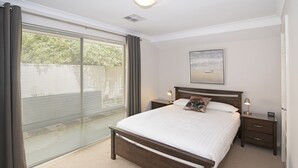 Un dormitorio con una cama grande, una mesita de noche, una lámpara de noche, una ventana con persianas y un cuadro en la pared.