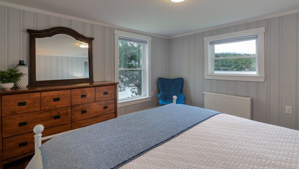A bedroom with a wooden dresser, a large mirror, a blue chair, a bed with a blue and white blanket, and two windows with views of greenery. - Lakefront home, water-view porch (Dedham)