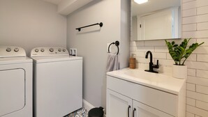 A laundry room with two white washing machines, a towel rack, a sink with a black faucet, and a potted plant. - Lakefront home, water-view porch (Dedham)