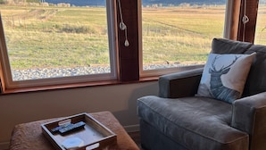 A living room with a large window offering a view of mountains and a field. There is a gray sofa, a wooden ottoman with a tray holding a remote control, and a pair of shoes on the floor.