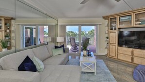 Un salon avec un canapé, une table basse et un meuble pour la télévision. Il y a un balcon avec vue sur l’océan et les palmiers.