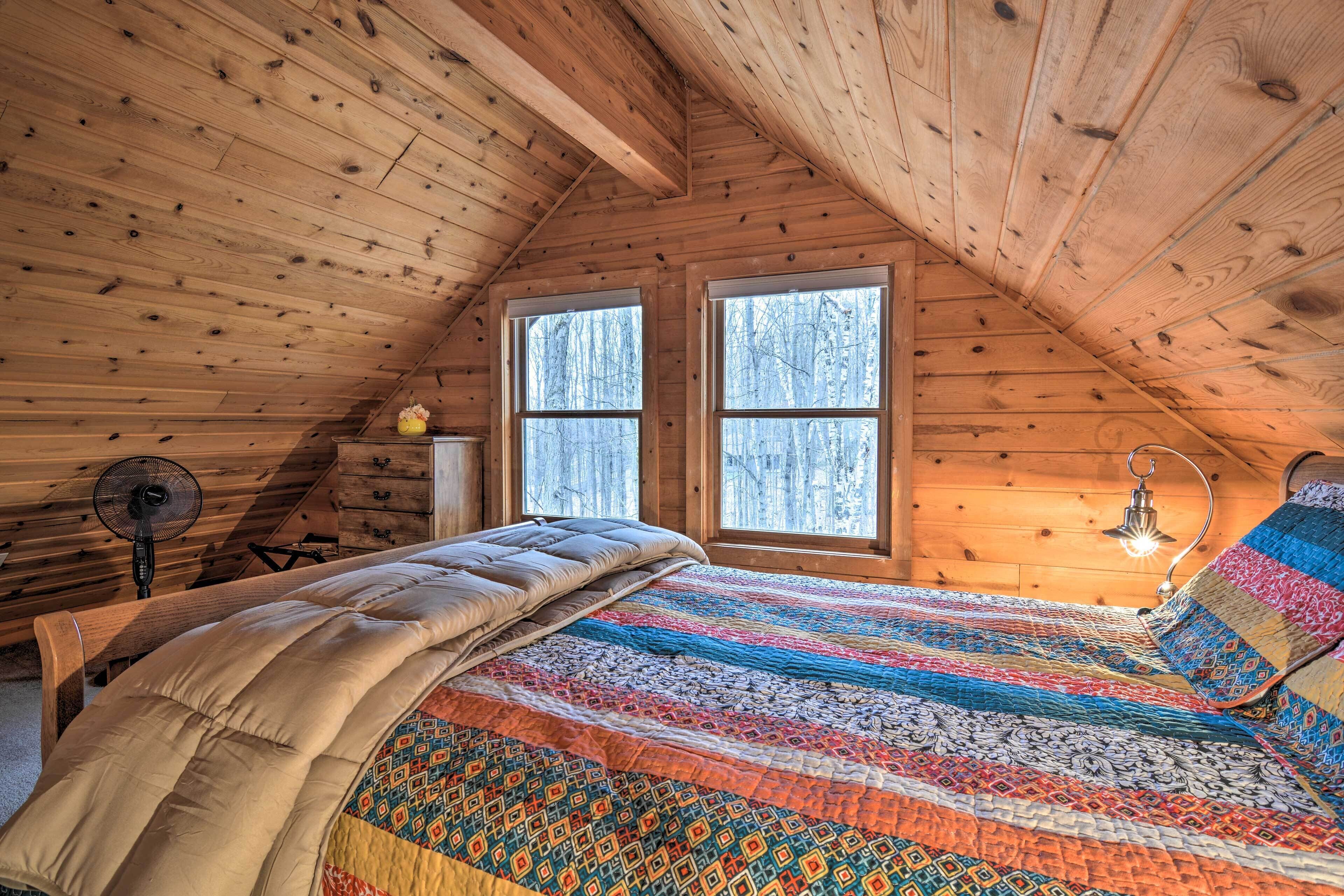 A cozy attic bedroom with a wooden ceiling, a large bed with a colorful quilt, two windows, a fan, and a bedside lamp.