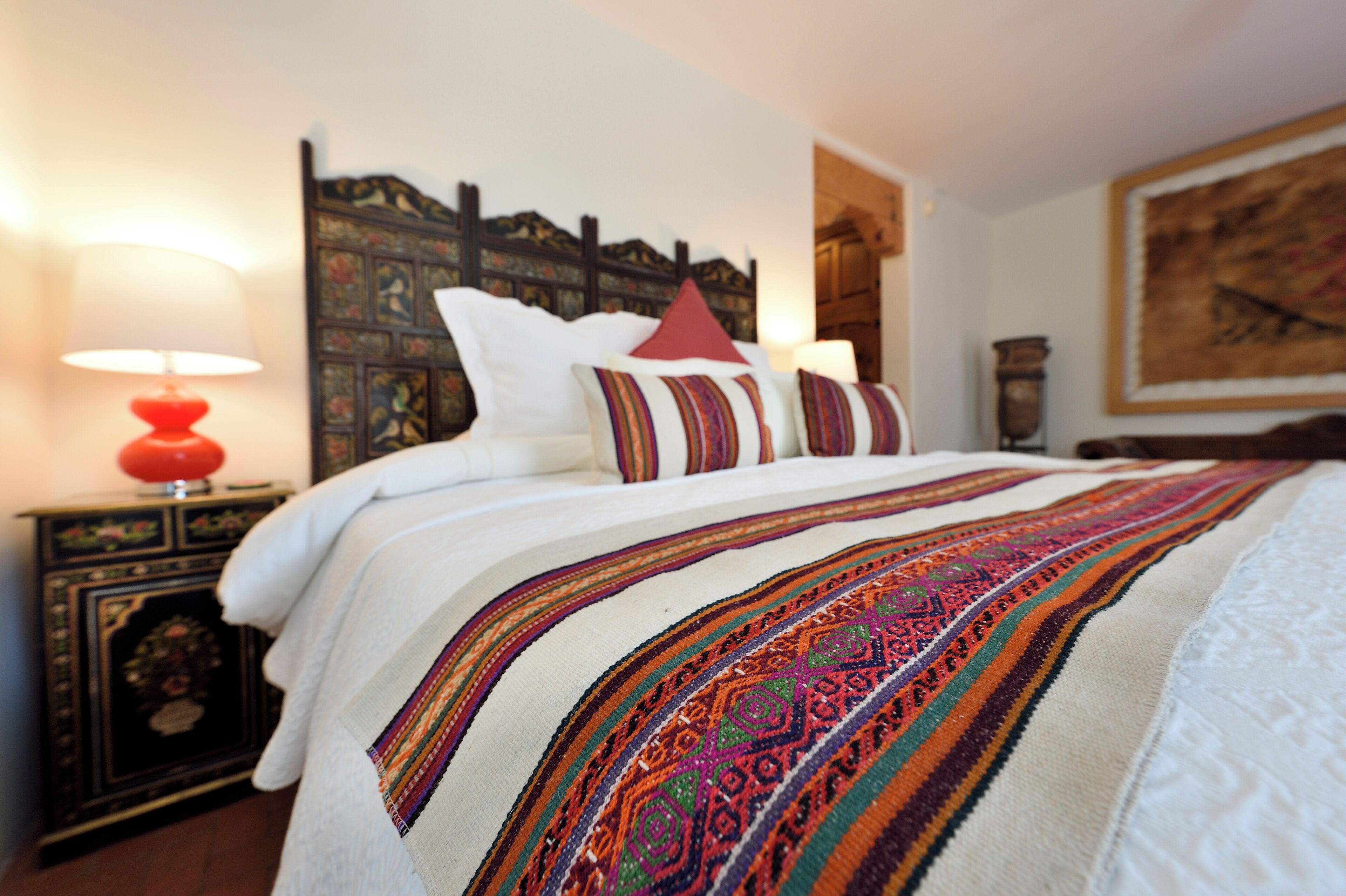 A neatly made bed with a patterned blanket, a red lamp, and a decorative headboard.