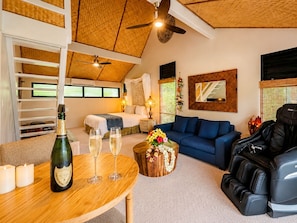 Ein Zimmer mit einem Bett, einem blauen Sofa, einem Massagesessel, einem Holztisch mit Flasche und Gläsern sowie einem Deckenventilator.