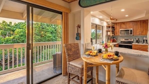 Un balcón con vistas a exuberante vegetación, una mesa de comedor con frutas y bebidas, y una cocina con armarios de madera y electrodomésticos modernos.