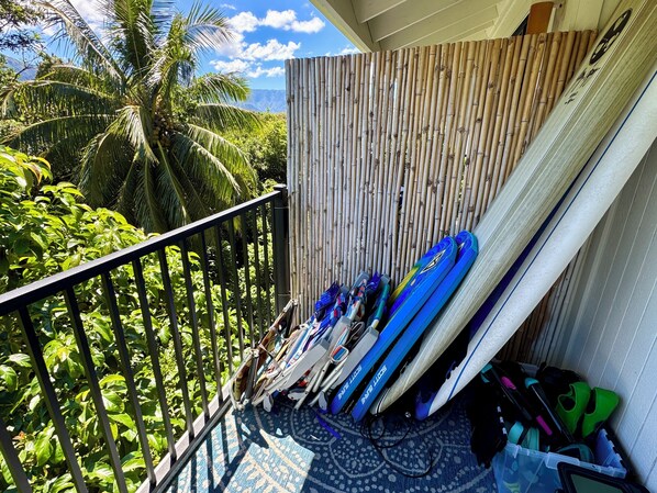 Un balcón con vistas a vegetación exuberante y montañas, cerca de una valla de bambú y con tablas de surf apoyadas en la pared.