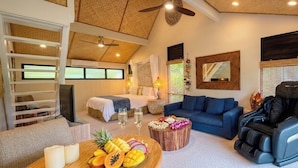 Amplia sala de estar con ventilador de techo, un sillón de masaje, un sofá azul, una mesa de centro de madera con fruta y una cama con dosel blanco.