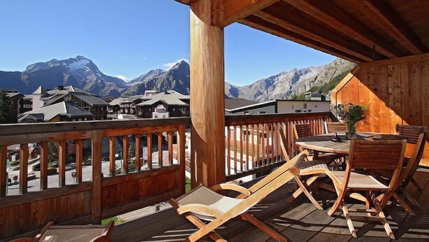 A wooden deck with a table and chairs, overlooking a village with traditional buildings and mountains in the background.