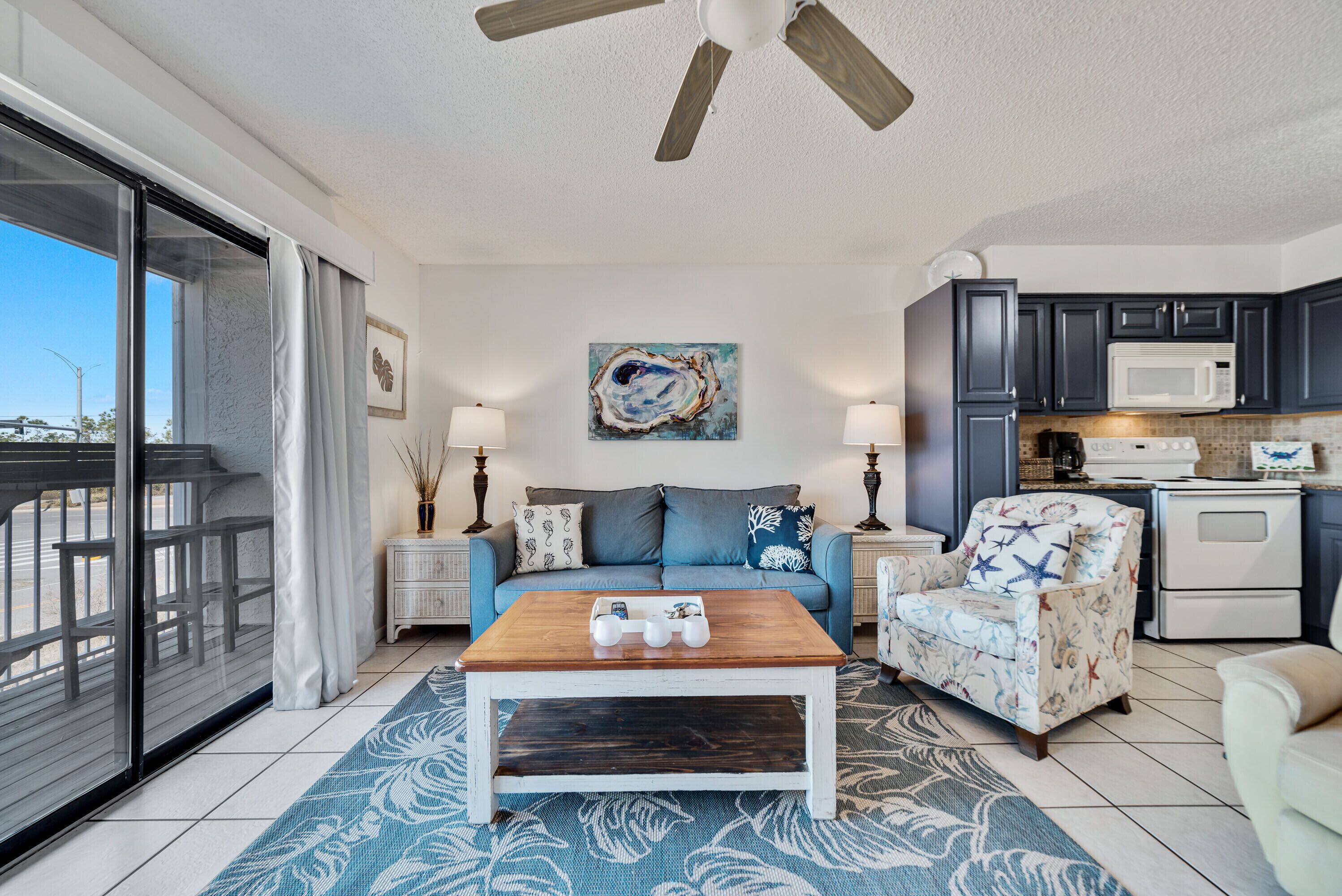 A living room with a wooden coffee table, a sofa, a patterned rug, a ceiling fan, and a kitchen area with dark cabinets and a microwave.