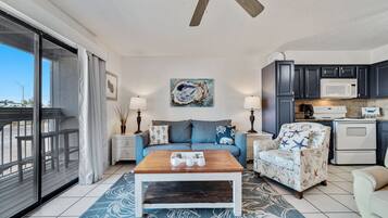 A living room with a wooden coffee table, a sofa, a patterned rug, a ceiling fan, and a kitchen area with dark cabinets and a microwave.
