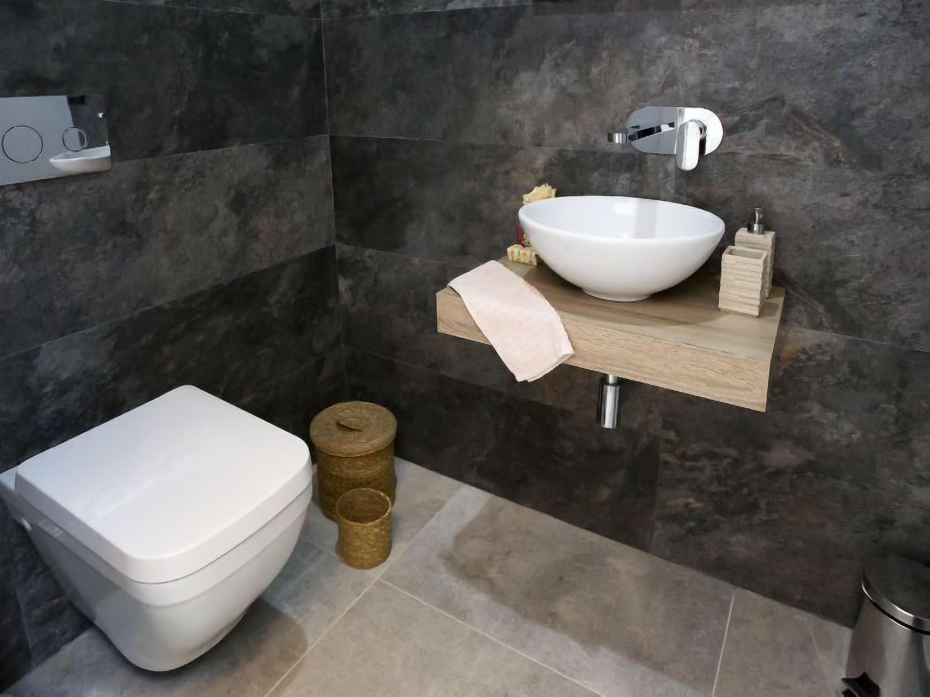 Et moderne badeværelse med et hvidt toilet, en hvid vask med en skål og en hylde under vasken i træ.