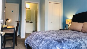 Une chambre à coucher avec un lit, un bureau avec une lampe, une chaise, et une salle de bain visible à travers une porte ouverte.