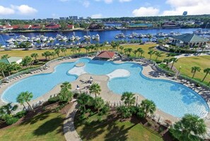 A large, uniquely shaped swimming pool surrounded by palm trees and lounge chairs, with a marina and buildings in the background.