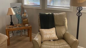 A beige leather armchair with a 'Beach House' pillow, a glass coffee table with decorative items, and a lamp. - Myrtle Beach Resort Unit 5110 - Ideal Beach Getaway (Myrtle Beach)
