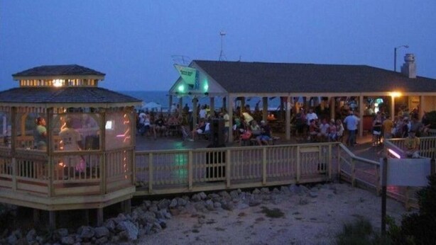 A beachside pavilion with a wooden deck and a glass enclosure, overlooking the beach.