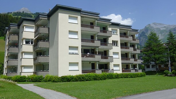 A multi-story apartment building with balconies, a paved driveway, and a mountainous backdrop.