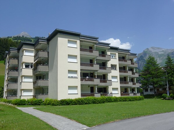 Un edificio de apartamentos de varios pisos con balcones, un camino de entrada pavimentado y montañas al fondo.