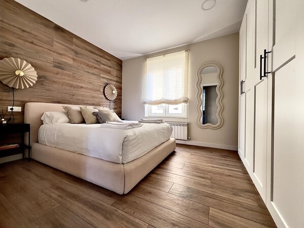 A bedroom with a wooden wall, a bed with white bedding, a bedside table, a mirror, and a window with blinds. - Housingleón - Muralla II (León)