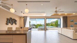 A spacious living area with a large glass door leading to a pool, a ceiling fan, and a dining area with a glass table. - Teetara Villa Patong (Patong)