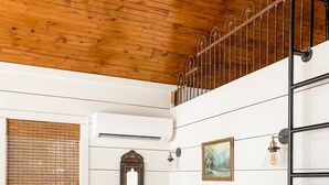 Un dormitorio con techo de madera, una cama con ropa blanca, cabecera de madera, un taburete pequeño y una estantería en la pared.