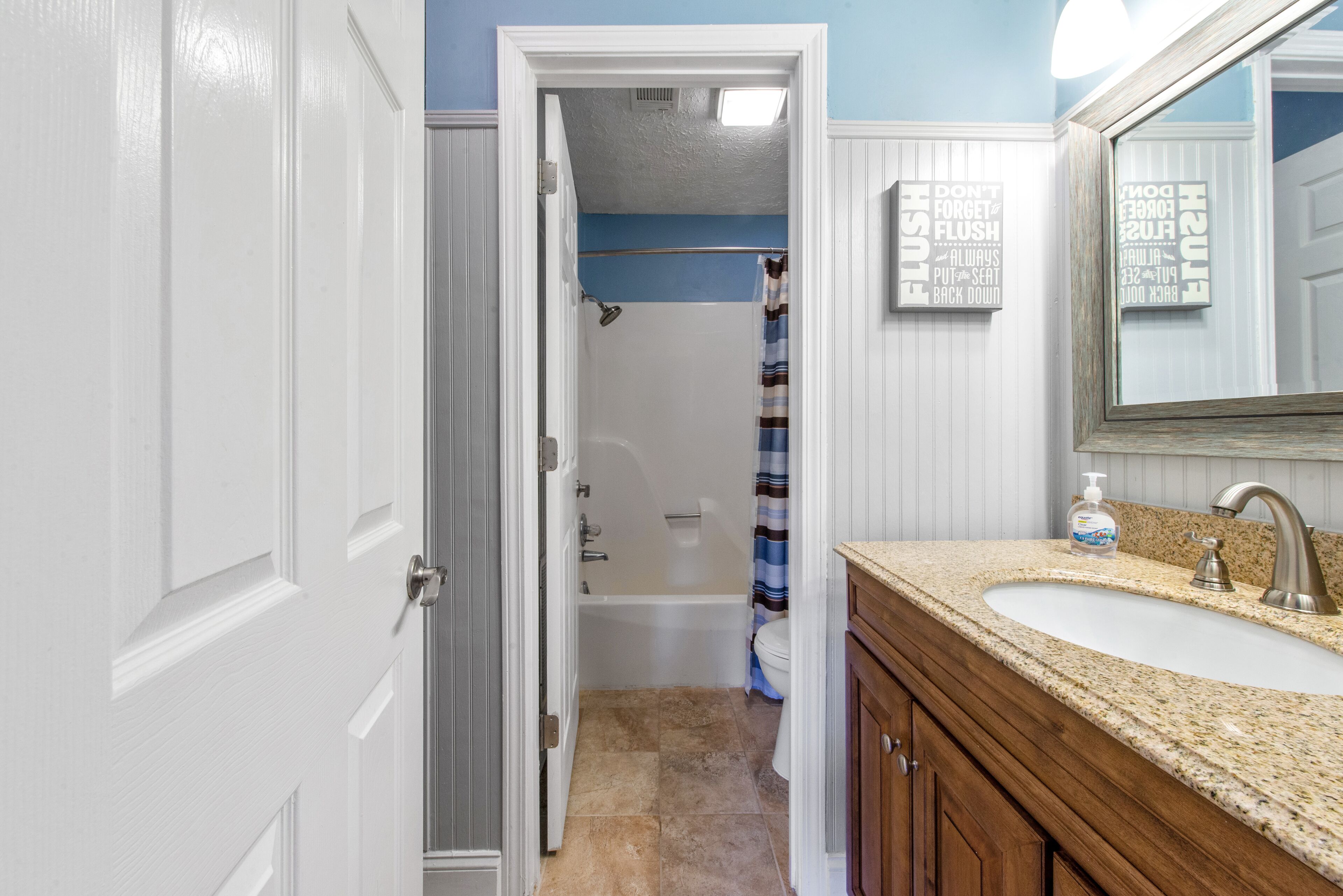A bathroom with a white door, a shower with a striped curtain, a sink with a soap dispenser, and a mirror with a sign above it.