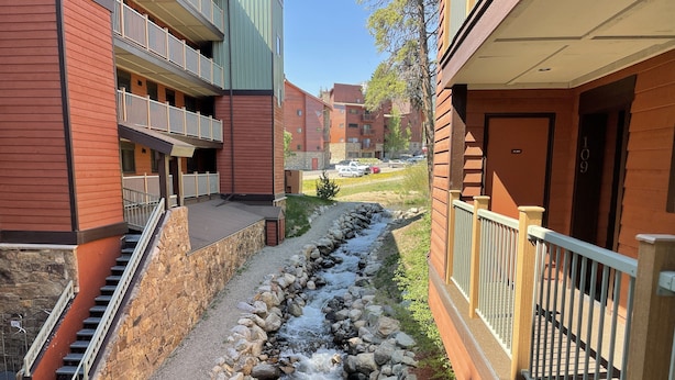 A residential building with a stone staircase leading to a balcony, a small stream running through a landscaped area, and a clear view of the street in the background. - Perfect location off 4 o'clock run, Walk to Mainstreet, Ski-In, Shared Hot Tub (Breckenridge)