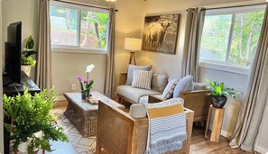 A living room with a sofa, armchairs, a TV, and a lamp. There are plants and a painting on the walls.