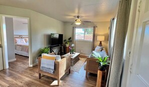 A living room with a television, wicker furniture, a ceiling fan, and a window with curtains. - Sweet Country Cottage (Kilauea)