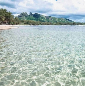 Uma praia com águas claras e rasas, um areal e uma cadeia de montanhas ao fundo.