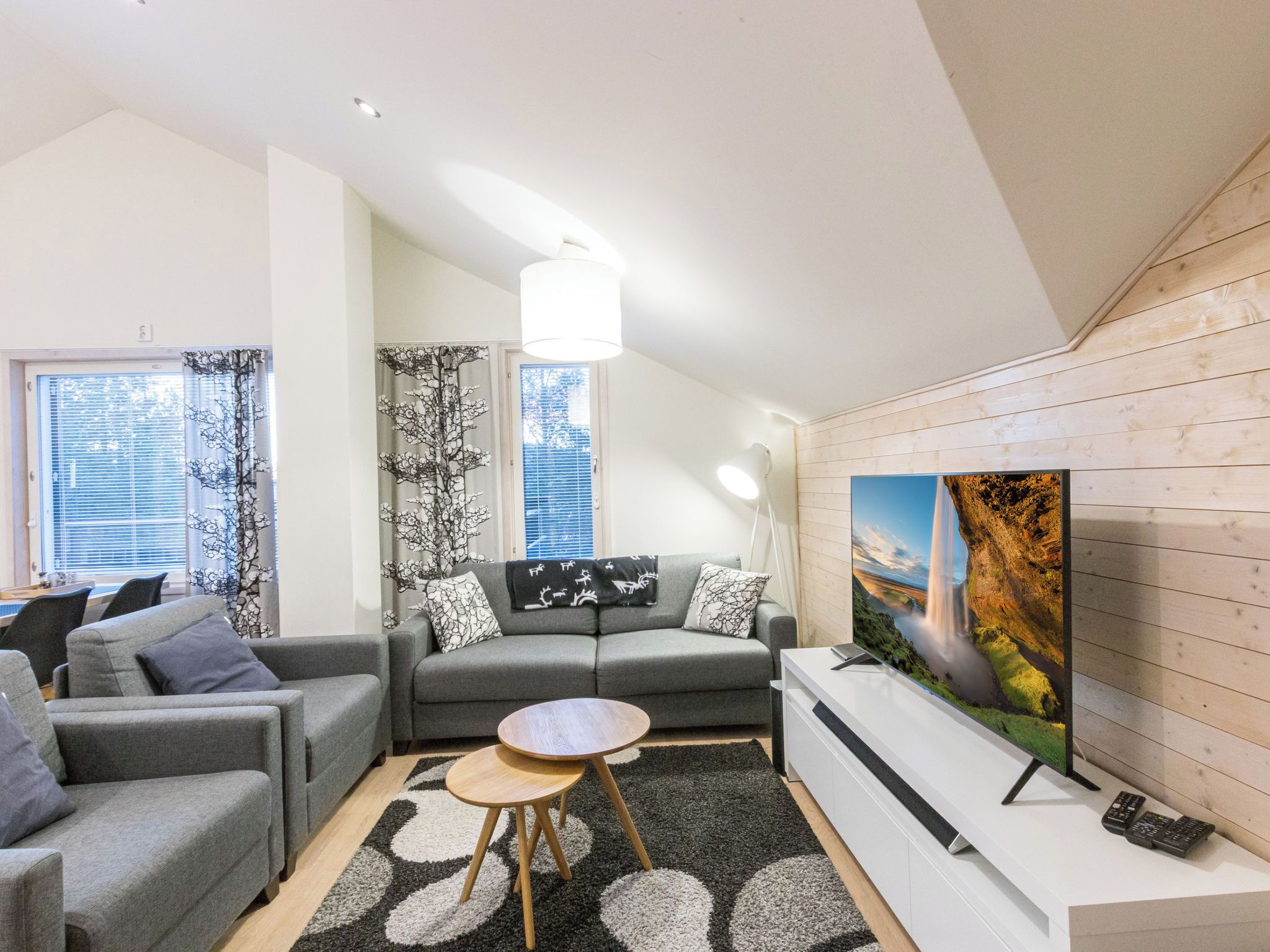 A modern living room with a grey sofa, a round wooden coffee table, and a flat-screen TV mounted on a wooden wall.