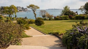 Un jardín costero con un sendero pavimentado que lleva a una zona verde, con vistas a una masa de agua con barcos y un paisaje urbano lejano.