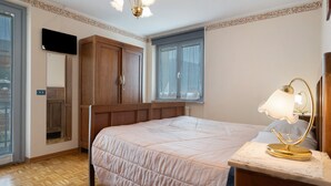 A bedroom with a bed, a nightstand with a lamp, a wardrobe, a mirror, and a window with curtains.