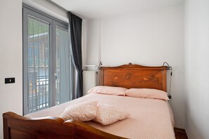 A bedroom with a wooden bed, a large window with curtains, and a view of a balcony. - Vacation Apartment Ciclamino Standard Vittoria with Mountain View, Wi-Fi & Garden (Dorsino)