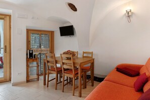 A dining area with a wooden table and chairs, a TV mounted on the wall, and a window with patterned curtains.