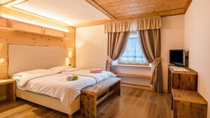 Una camera da letto con soffitto in legno, un letto con biancheria bianca, una panca, una TV su un mobile in legno e una finestra con tende.