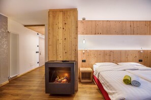 A modern bedroom with a built-in fireplace, wooden panel walls, a bed with white linens, and a small bedside table. - Chalet "Osmar Suite 1" with Mountain View, Balcony & Wi-Fi (Campitello di Fassa)