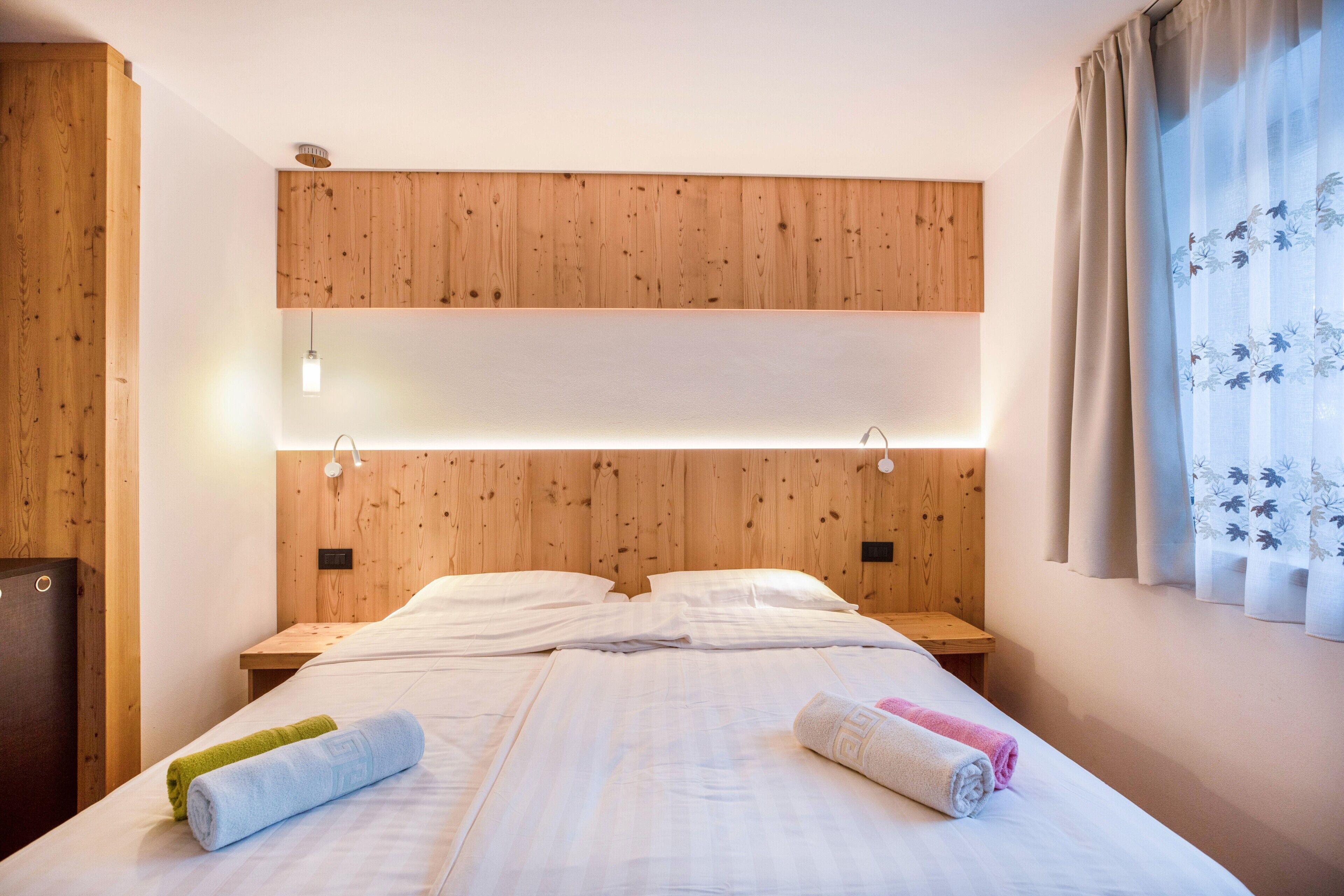 Een hotelkamer met een houten hoofdbord, een bed met witte lakens, twee nachtkastjes en een raam met gordijnen.