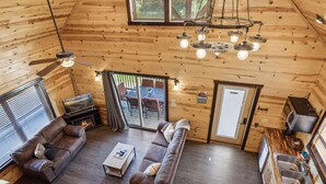 A cozy cabin interior with a wooden ceiling, a sofa set, a dining table, a fireplace, and a kitchen area.